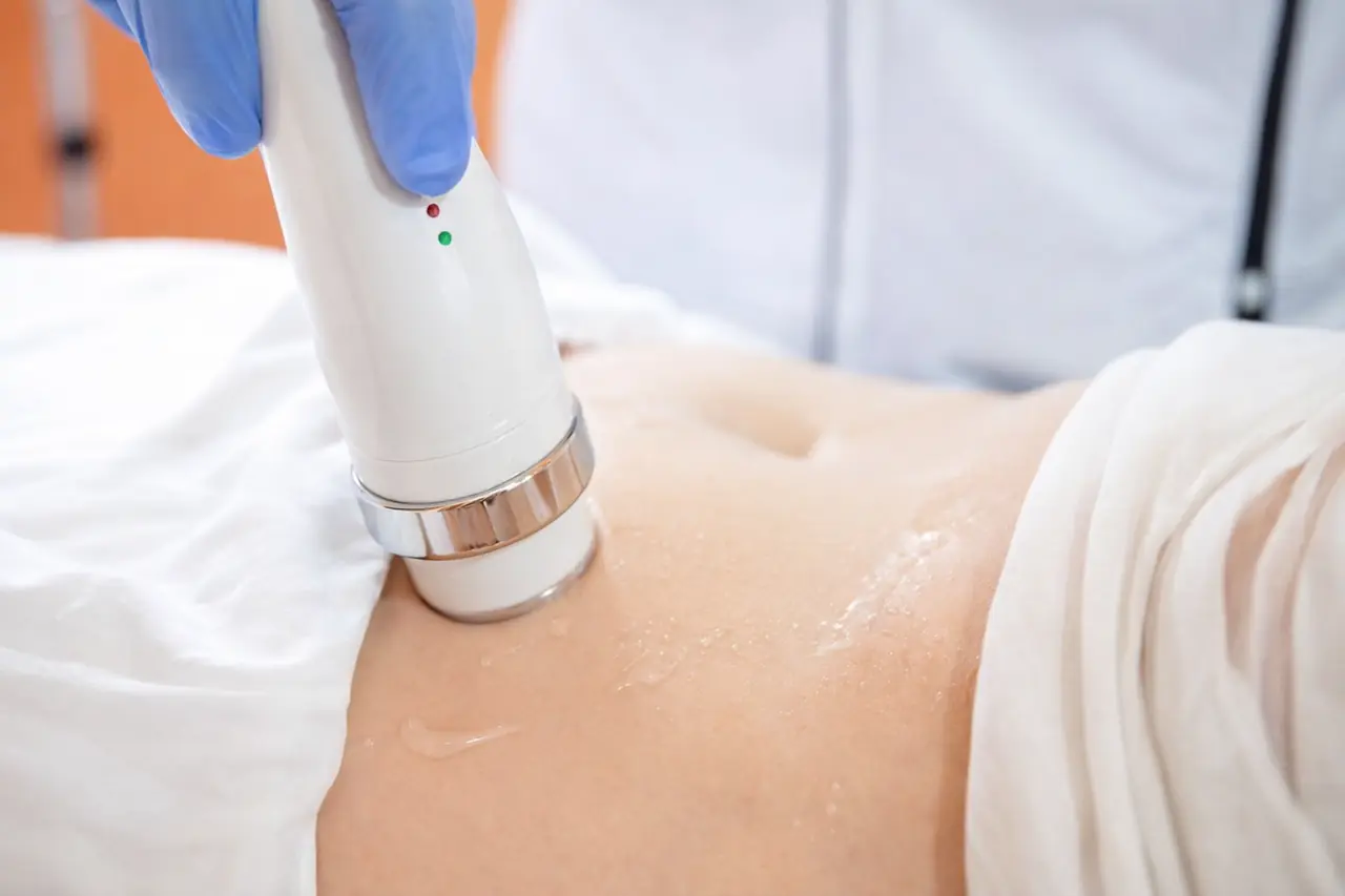 Gloved hand guiding a white handheld device over a gel-coated abdomen during an EMS body sculpting session