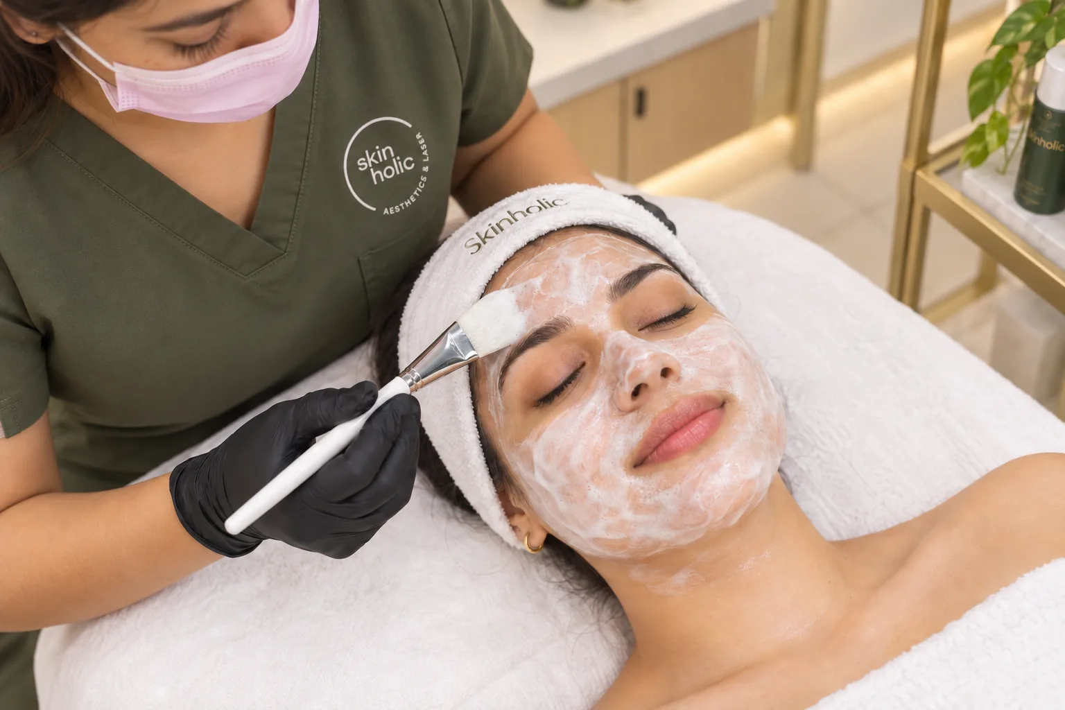 Masked practitioner brushes a white cream mask onto a smiling woman's face during a brightening facial