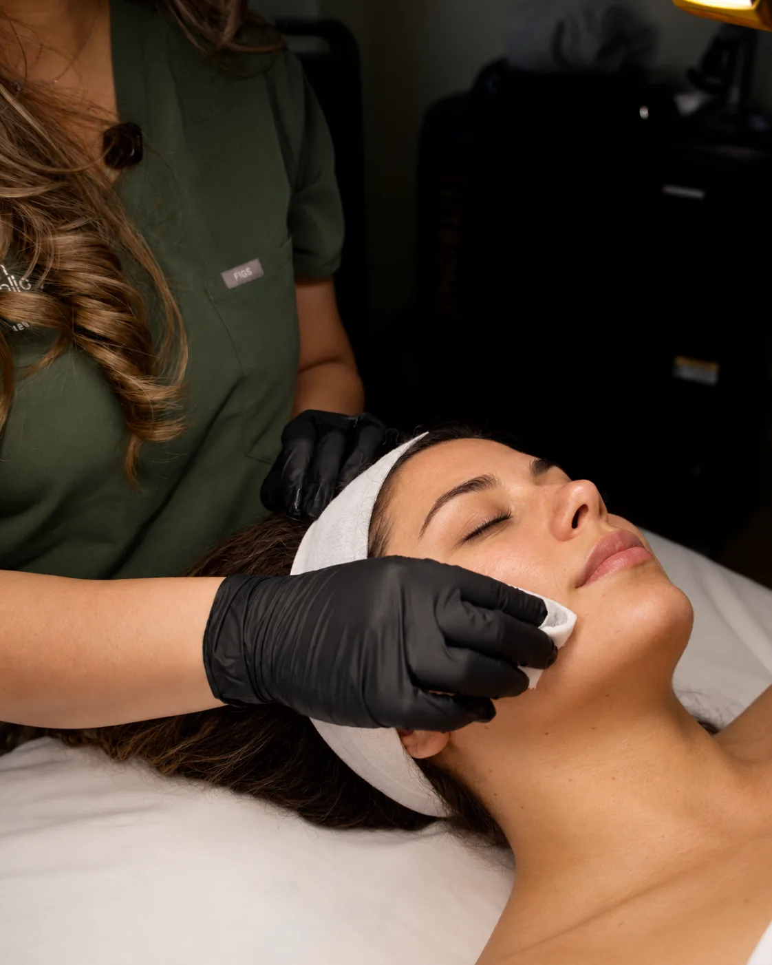 Gloved esthetician presses a cotton pad to the lower cheek of a woman in a headband, eyes closed, during a chemical peel