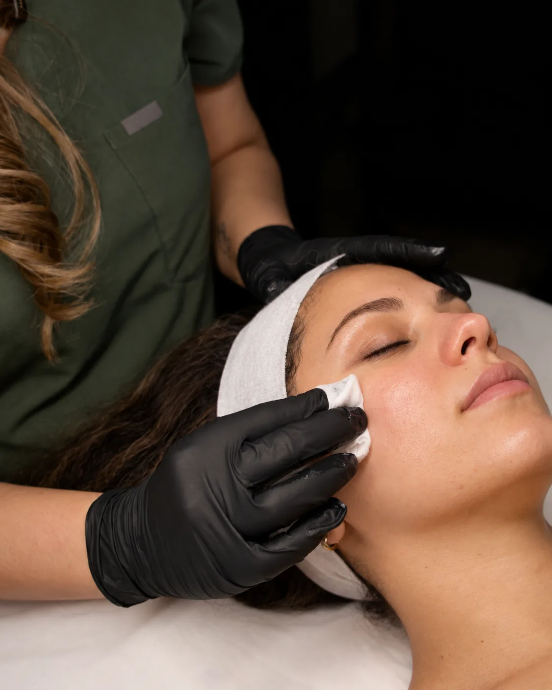 Close view of gloved hands holding a cotton pad to a woman's cheek and steadying her forehead during a chemical peel