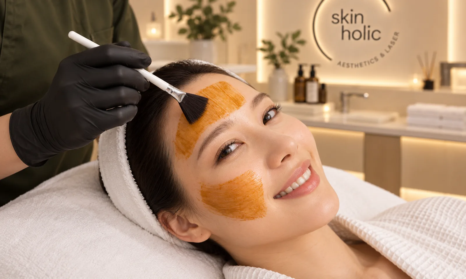 Smiling woman reclines as a gloved practitioner brushes an orange Cosmelan mask onto her forehead and cheek