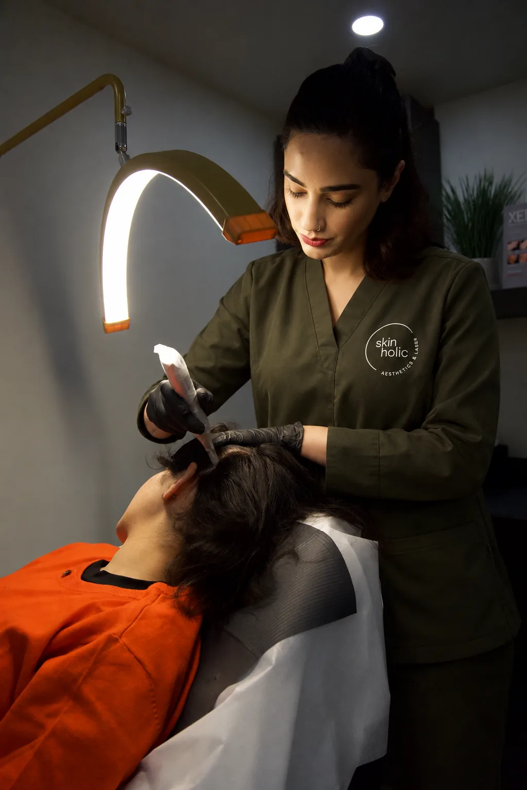 Practitioner under an arc lamp performs scalp microneedling on a reclined person in a dim room, DERIVE hair wellness