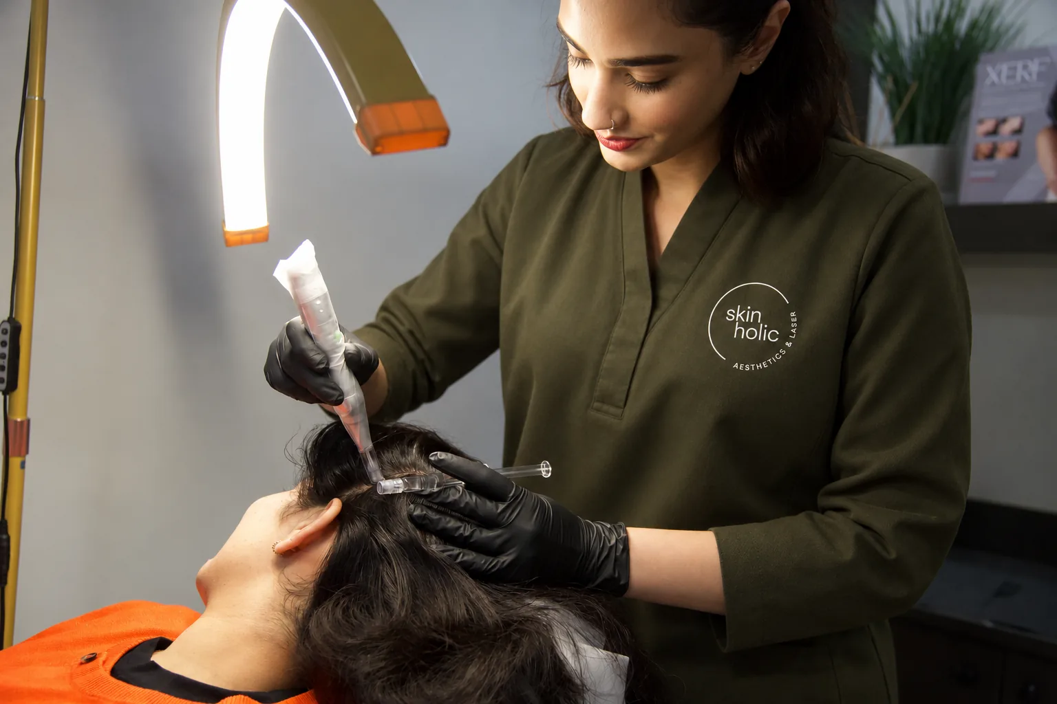Practitioner in an olive top uses a microneedling pen on a reclined person's scalp under an arc lamp, DERIVE hair wellness