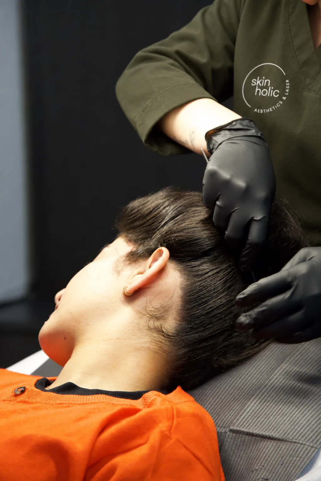 Gloved practitioner in an olive top parts the dark hair of a reclined person to expose the scalp for DERIVE hair wellness