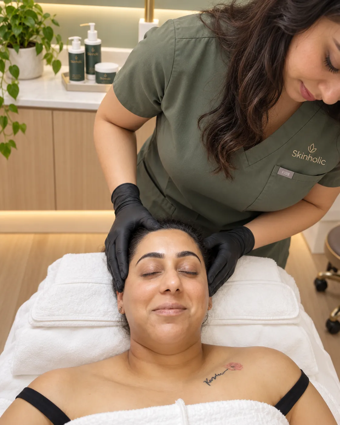Practitioner in olive scrubs and black gloves cradles a smiling woman's head on a treatment bed before dermaplaning