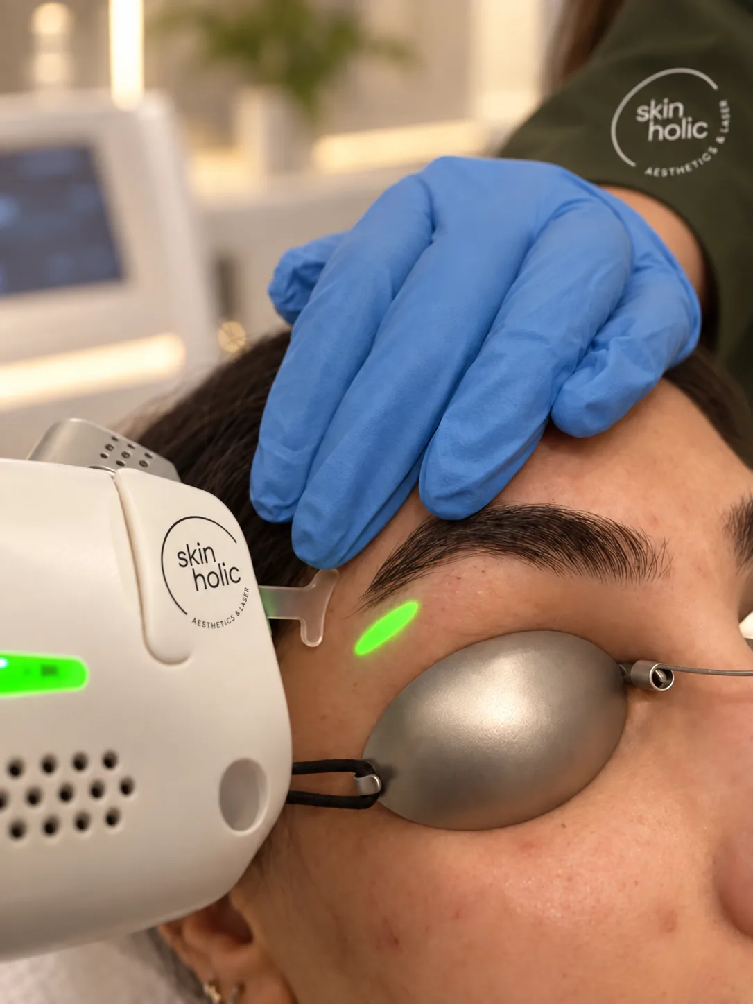 Gloved hand steadies a woman's brow as a pico laser handpiece casts a green aiming light beside her metal eye shield