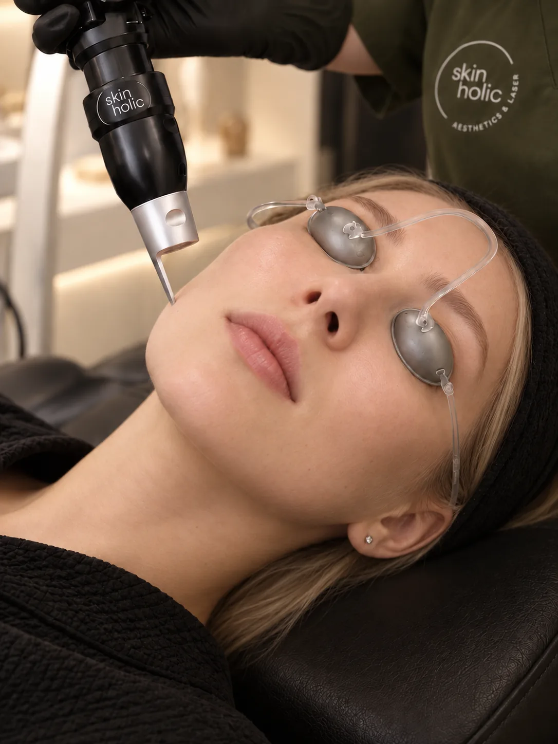 Woman wearing metal eye shields reclines as a slim laser handpiece is held near her lower cheek for Pico Laser Rejuvenation