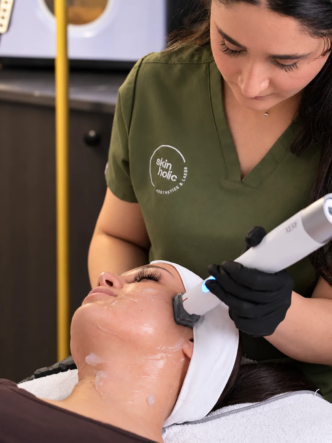 Practitioner in olive scrubs guides a skin tightening handpiece over gel on a woman's upper cheek near the temple