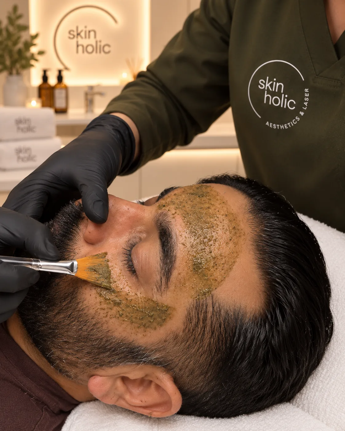 Close-up of gloved hands steadying a bearded man's face as a brush spreads green paste for a Zena Algae Peel